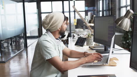 Junger-Asiatischer-Mann-Arbeitet-In-Einem-Modernen-Büroumfeld-Am-Laptop-Und-Trägt-Kopfhörer-Und-Eine-Mütze