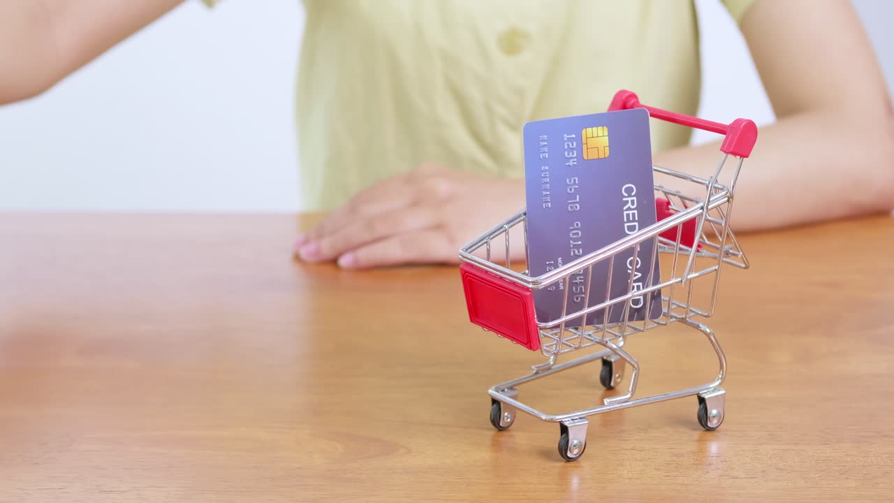 A hand places a credit card into a miniature shopping cart symbolizing online shopping and consumer transactions