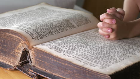 young child reading bible with hands crossed on top of book, close up view