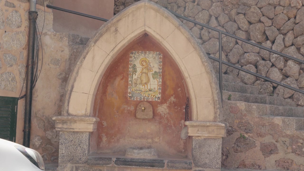 Una nicchia di fontana di paese ad arco gotico decorata con piastrelle di ceramica raffiguranti un santo pastore. Una scala in pietra sale dietro il muro consumato dalle intemperie. Inquadratura media