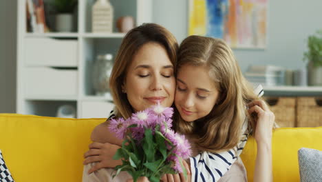 Close-Up-Of-The-Beautiful-Mother-And-Daughter-On-The-Sofa-Hugging-And-Woman-Holding-A-Bouquet-Of-Flowers-Her-Daughter-Gave-Her