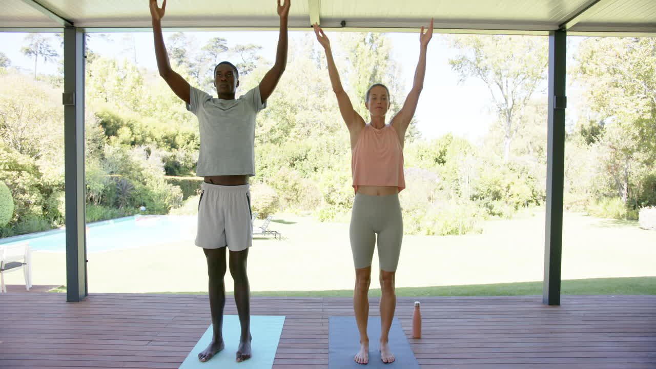 Diverse couple practicing yoga outdoors, raising arms up, at home