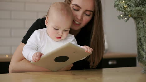 Mom-and-and-baby-sitting-at-the-table-playing-tablet-computer-pressing-your-fingers-on-the-screen-and-laughing