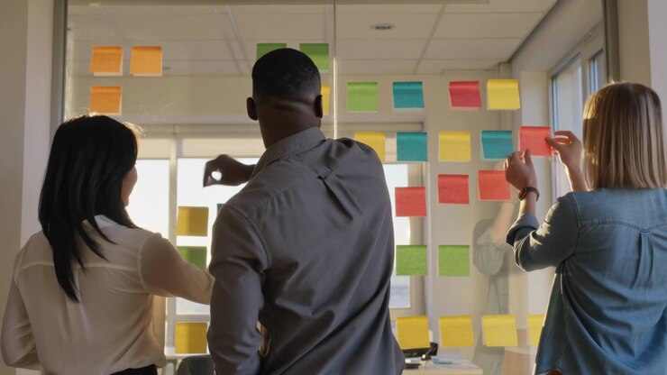 Un team di tre persone in abbigliamento business-casual inizia una riunione, posizionando post-it sul vetro per la pianificazione.