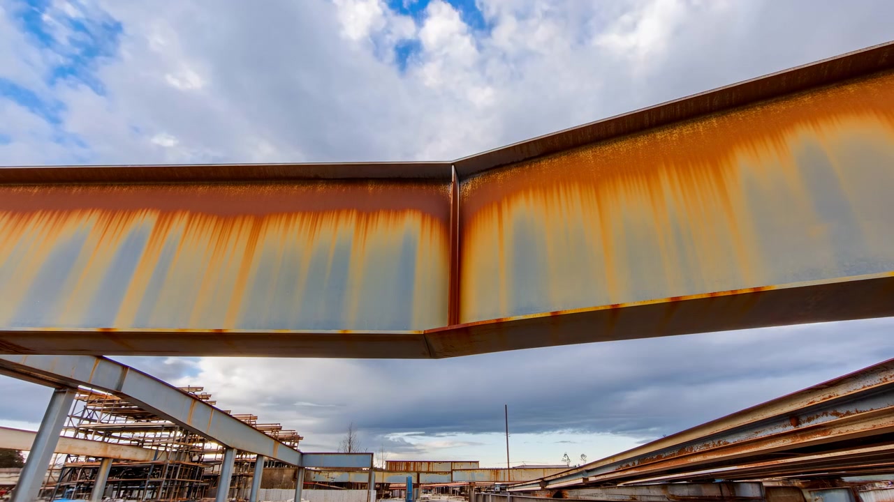 Des poutres métalliques patinées et des éléments structurels s'étendent sur le cadre supérieur, sur un ciel bleu clair, avec des matériaux de construction et une structure industrielle visibles.