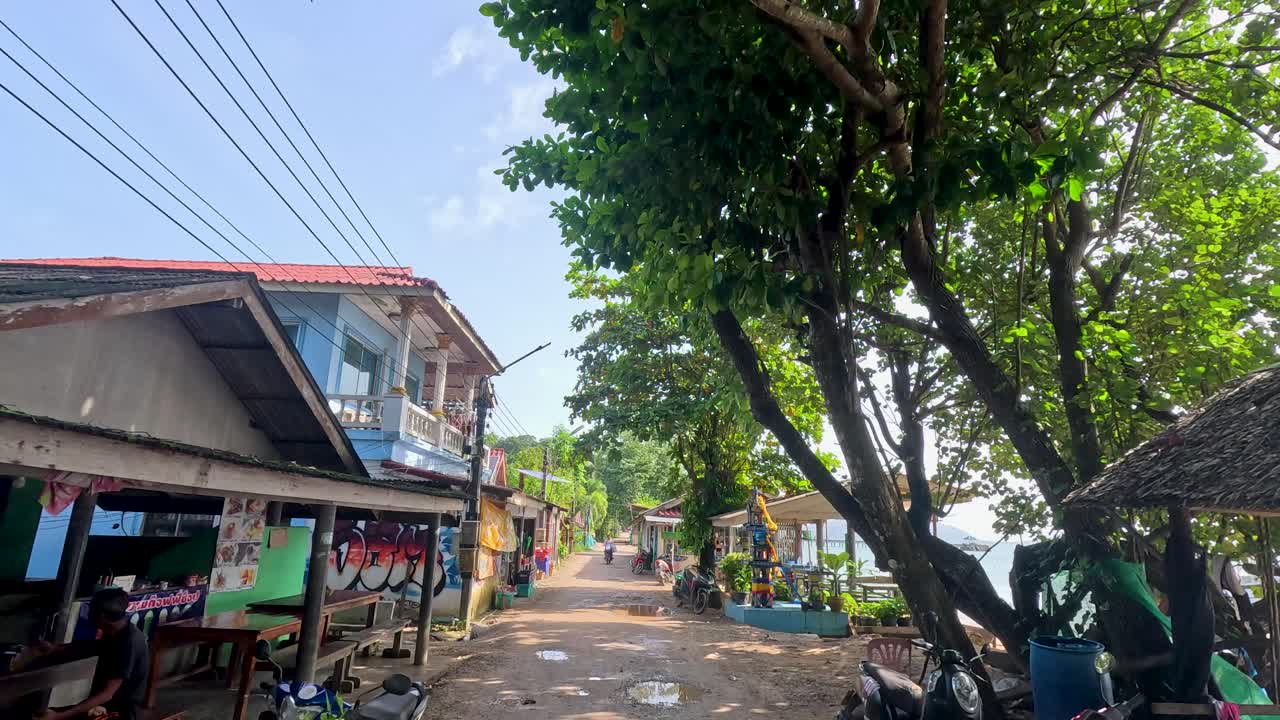 A handheld camera moves down a sunlit, tree-lined street in a coastal village, passing rustic shops and houses near the sea on Ko Phayam
