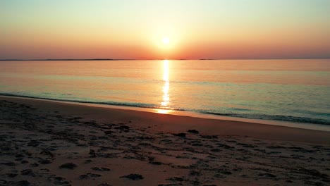 Luftaufnahme-Eines-Wunderschönen-Sonnenaufgangs-Am-Strand-Mit-Einer-Hellen,-Leuchtenden-Sonne,-Die-Farbenfrohe-Rote,-Orange,-Violette-Und-Gelbe-Reflexe-über-Friedliche,-Plätschernde-Wellen-Des-Meeres-Mit-Wunderschönem-Himmel-Und-Ozean-Wirft