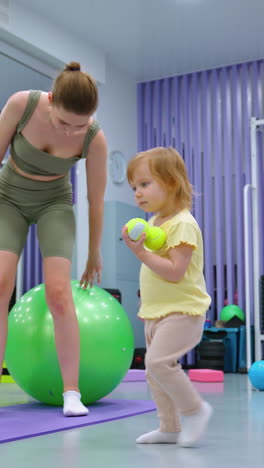 el niño recoge la pelota de tenis mientras que el adulto se sienta en la pelota verde de ejercicio en el gimnasio con decoración moderna, el entorno de fitness incluye equipos de entrenamiento, espejos y esteras de estiramiento