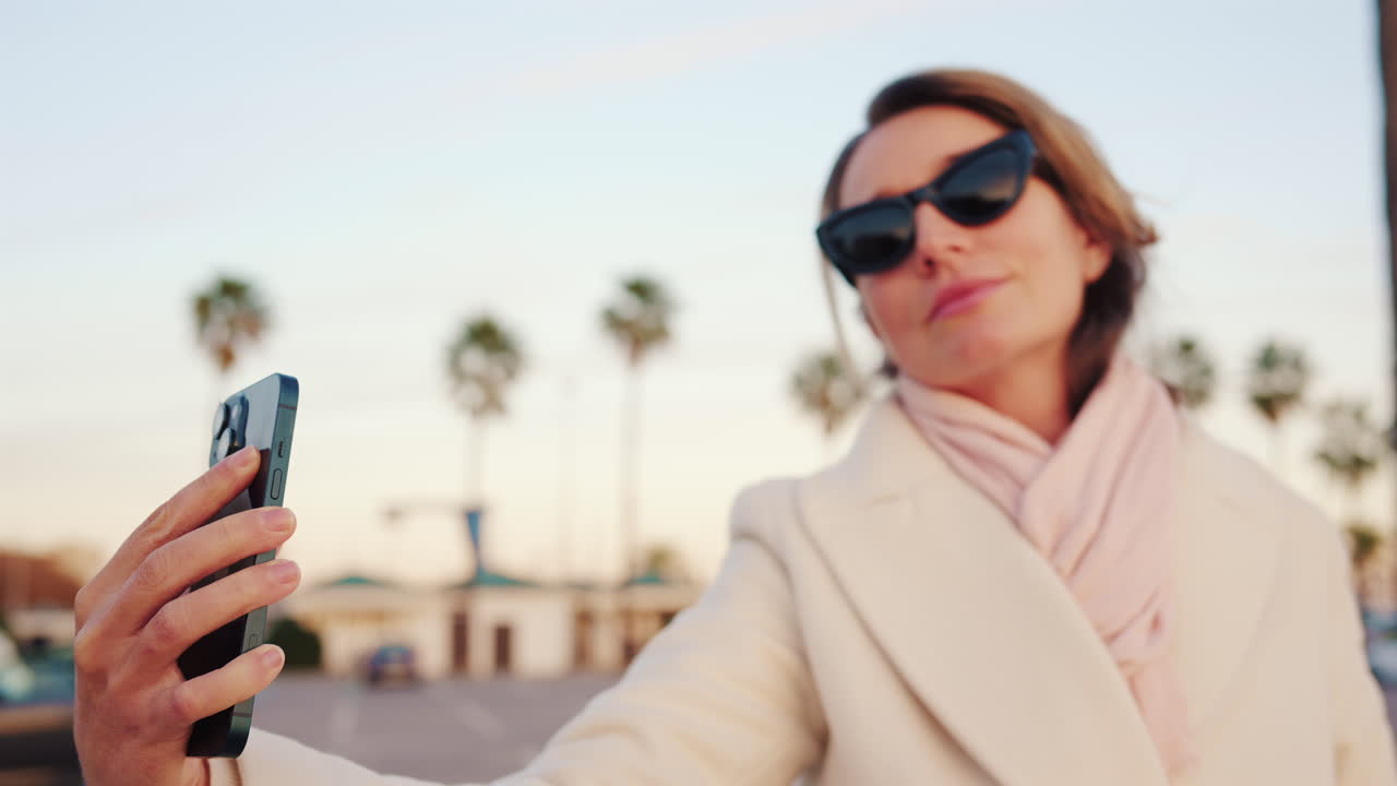Confident woman in a white coat takes a selfie with her smartphone in a sunny outdoor area lined with palm trees