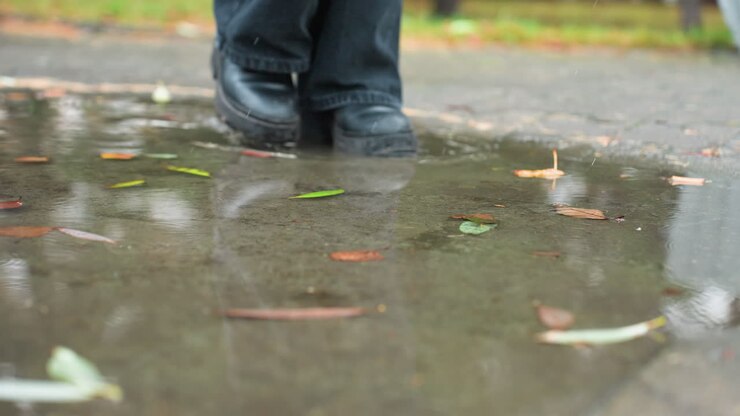 Primer plano de un charco de agua en un camino pavimentado y mojado con hojas de otoño de colores dispersas que reflejan los árboles desnudos de arriba, mientras una pierna con una bota negra pisa suavemente cerca durante un día nublado y tranquilo