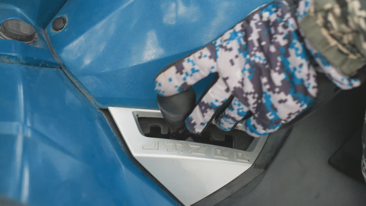 Primo piano della leva del cambio di un ATV, con una mano guantata che seleziona la marcia su un pannello di plastica blu. Un guanto mimetico afferra il pomello del cambio, con messa a fuoco ravvicinata sulla fessura meccanica. Si notano graffi di sporco e una texture ruvida, in preparazione per un'uscita off-road.