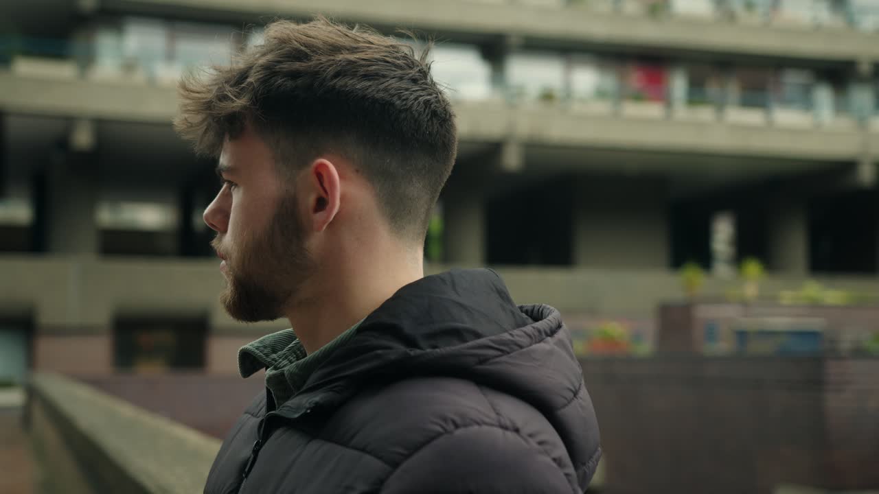 Side view of young man leaning up against wall in Barbican Estate with urban architecture behind him, staring out, shallow depth of field