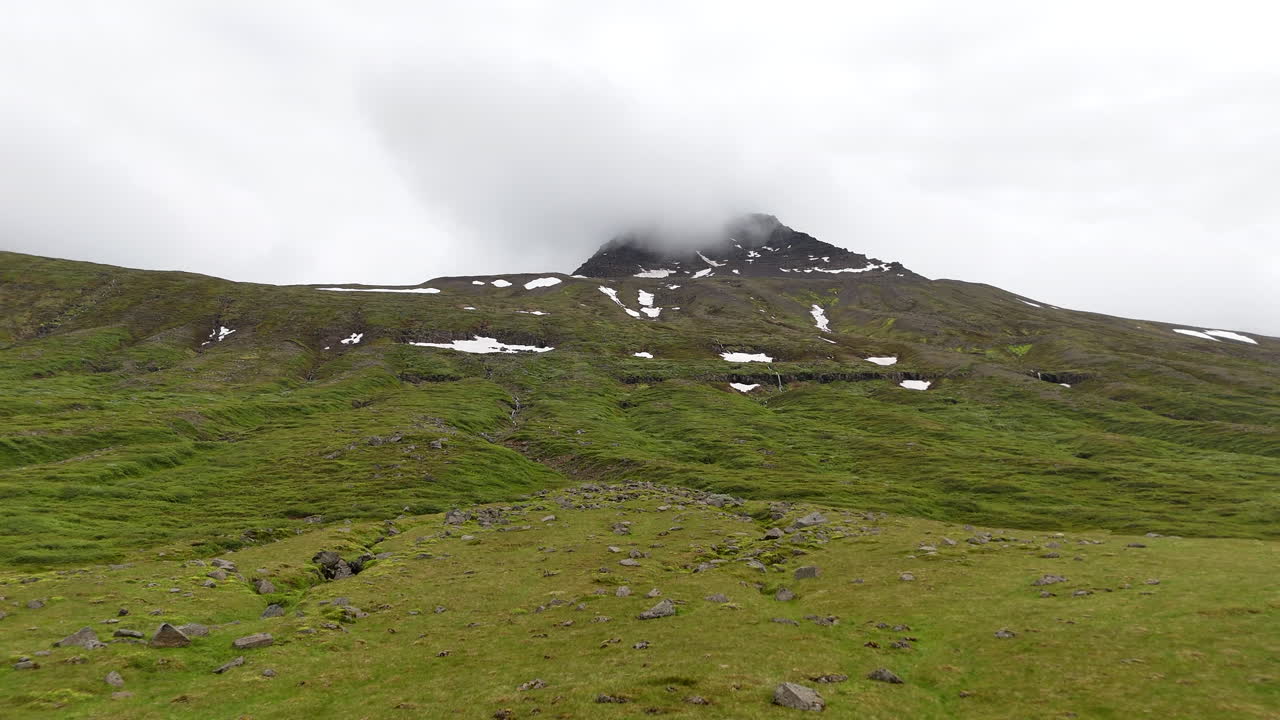 Wide aerial view of the mountains near Fáskrúðsfjörður, with snow-lined peaks, scattered waterfalls, winding wetlands, and an open valley crossed by a curving road under cloudy skies