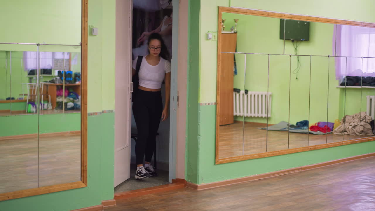 Young lady in casual outfit wearing white top and black leggings opens door, steps out gently and closes it while walking, large wall mirror shows her reflection inside bright indoor room