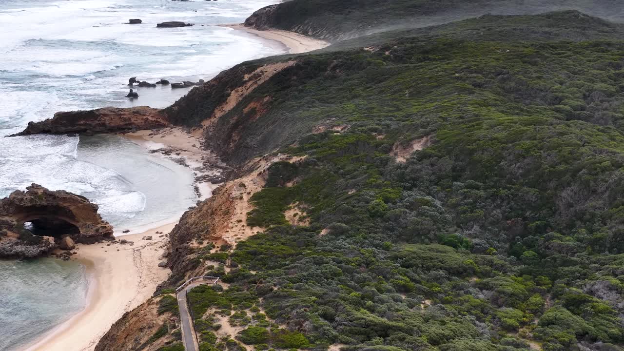 Drone glides above windswept cliffs, sandy beach, and ocean waves under overcast natural light