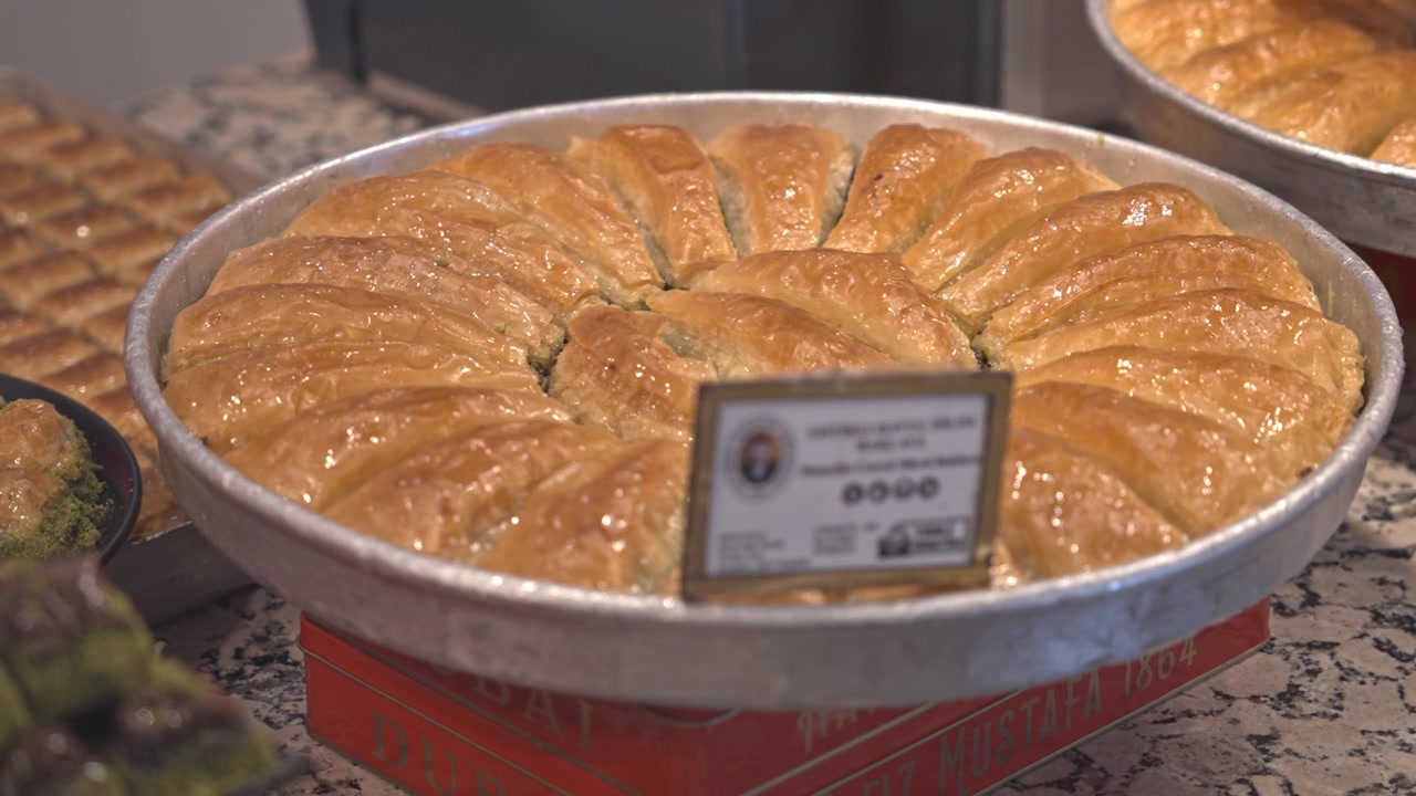 A mouth-watering 4K cinematic close-up of various traditional Turkish baklava types in a shop window. Features a red gift box labeled "ISTANBUL" among pistachio and walnut desserts.