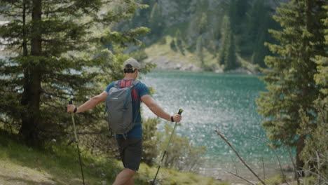 hiker walking with his hiking poles towards the mountain lake, path surrounded by conifers, water glistening