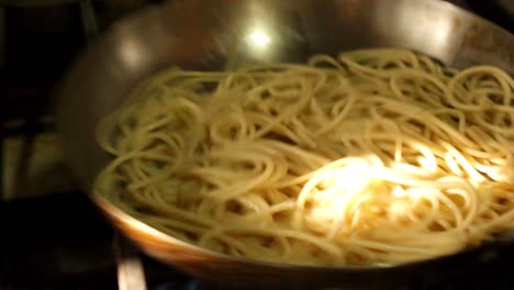 a chef's hand moving and mixing a metal pan with spaghetti on the fire