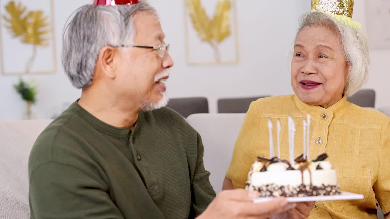 An elderly couple joyfully celebrates a birthday with cake, candles, and party hats in a warmly lit home setting