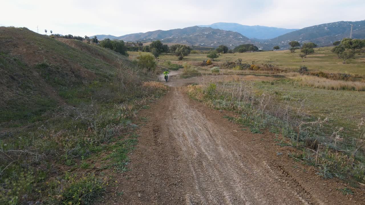 toma aérea de una pista de motocross rural en malaga españa con colinas, hierba y montañas en el fondo con una bicicleta de motocross saltando sobre una colina