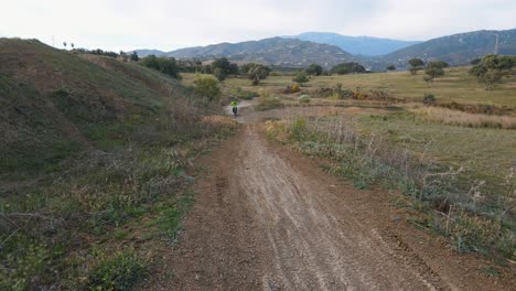 Aerial-dolly-shot-of-a-rural-motocross-track-in-Malaga-Spain-with-hills,-grass-and-mountains-in-the-background-with-a-riding-motocross-bike-jumping-over-a-hill