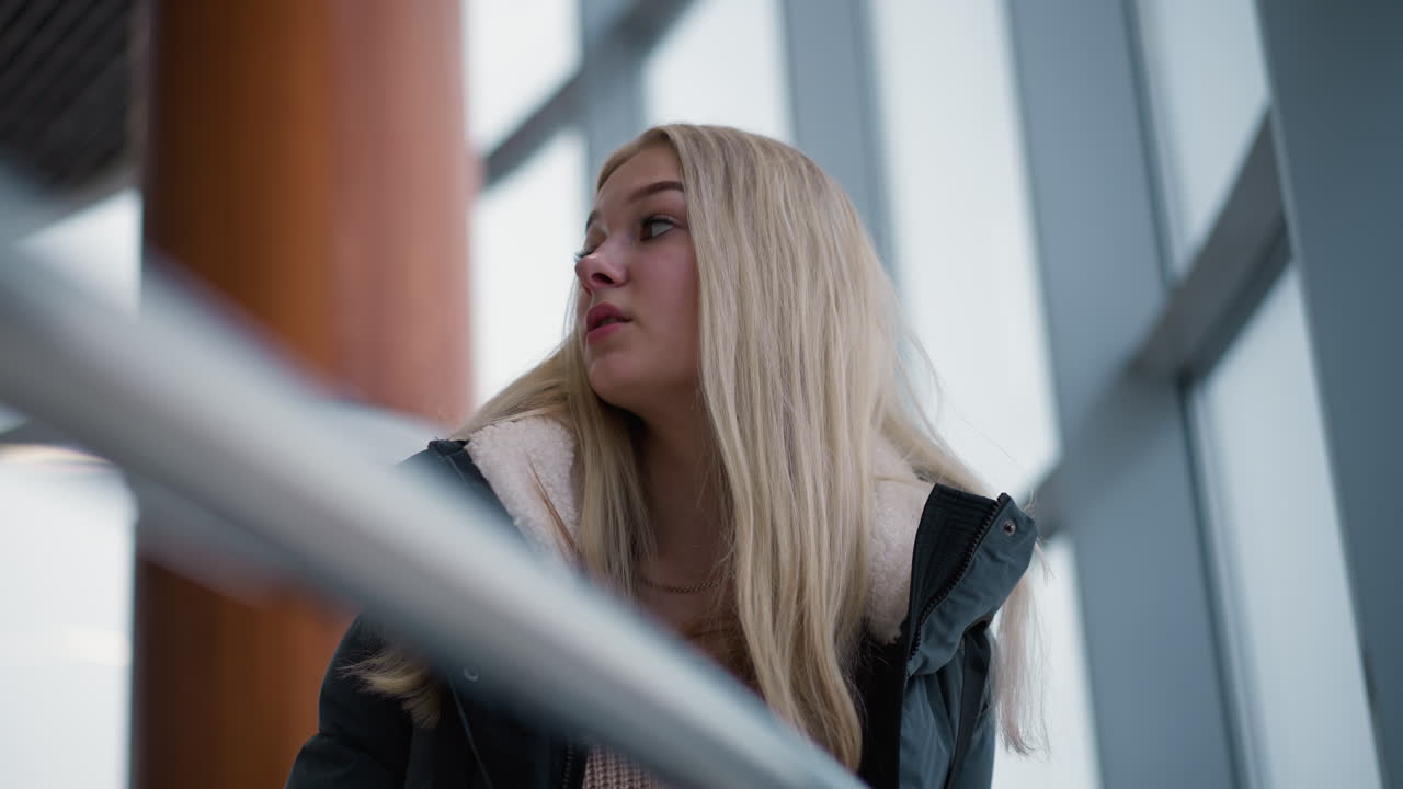 금발 머리카락과 따뜻한 미소를 가진 젊은 여인이 쇼핑몰에 서 있습니다. 그녀는 겨울 재을 입고 현대적이고 세련된 쇼핑 센터에 서서 유리 벽으로 둘러싸여있는 것처럼 만족합니다.