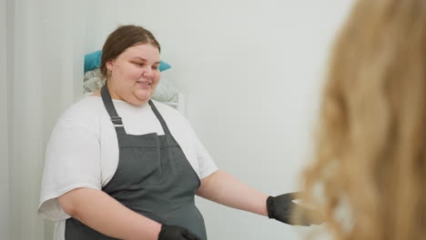client with long blonde hair walks into white lit salon and interacts with beautician standing near curtain. scene captures calm atmosphere and initial salon welcome in minimalist setting