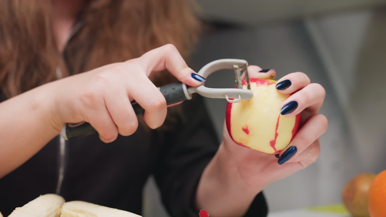 Student hand carefully peels red apple with handheld peeler in bright kitchen, blue nails visible, fruit prep for healthy snack, gradual smooth strokes, clean counter background