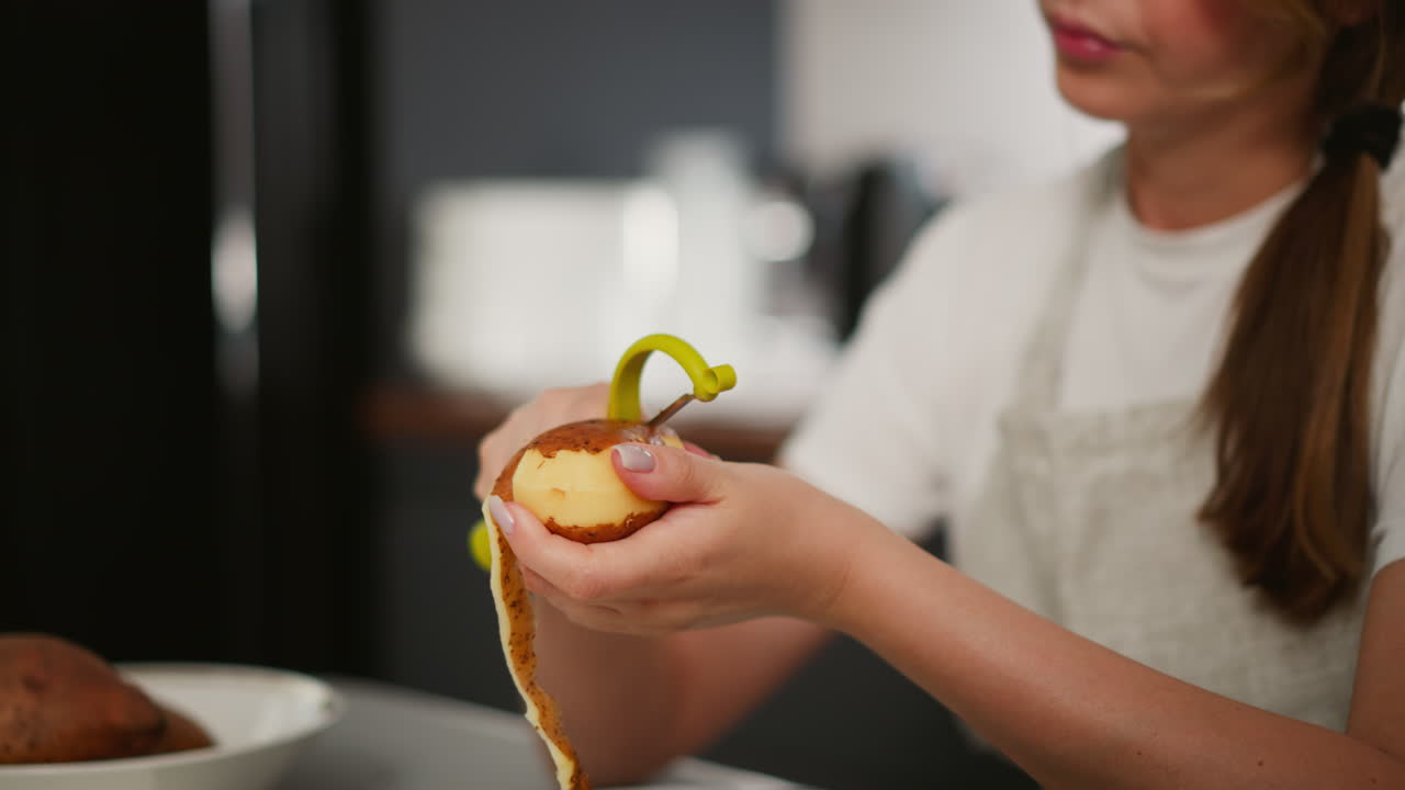 Close up of woman peeling raw potato using yellow peeler, long potato skin hanging from hand, focus on hands and potato, background softly blurred showing kitchen setup with bowl and appliances