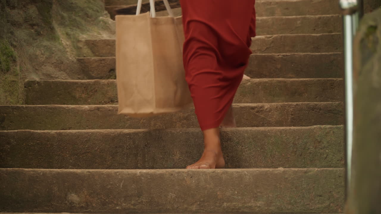 Graceful woman in scarlet dress steps quietly through lush garden. Serene scene of woman in crimson gown walking softly along stone stairs in peaceful garden surroundings with sunlight