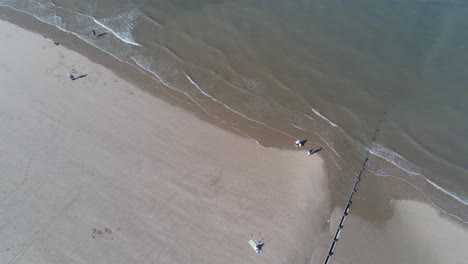 Soleada-Vista-Aérea-Mirando-Hacia-Abajo-Sobre-La-Playa-De-Arena-Dorada-Con-Turistas-Caminando-Debajo-De-La-Inclinación-Hacia-Arriba-A-Través-De-La-Marea-Del-Océano