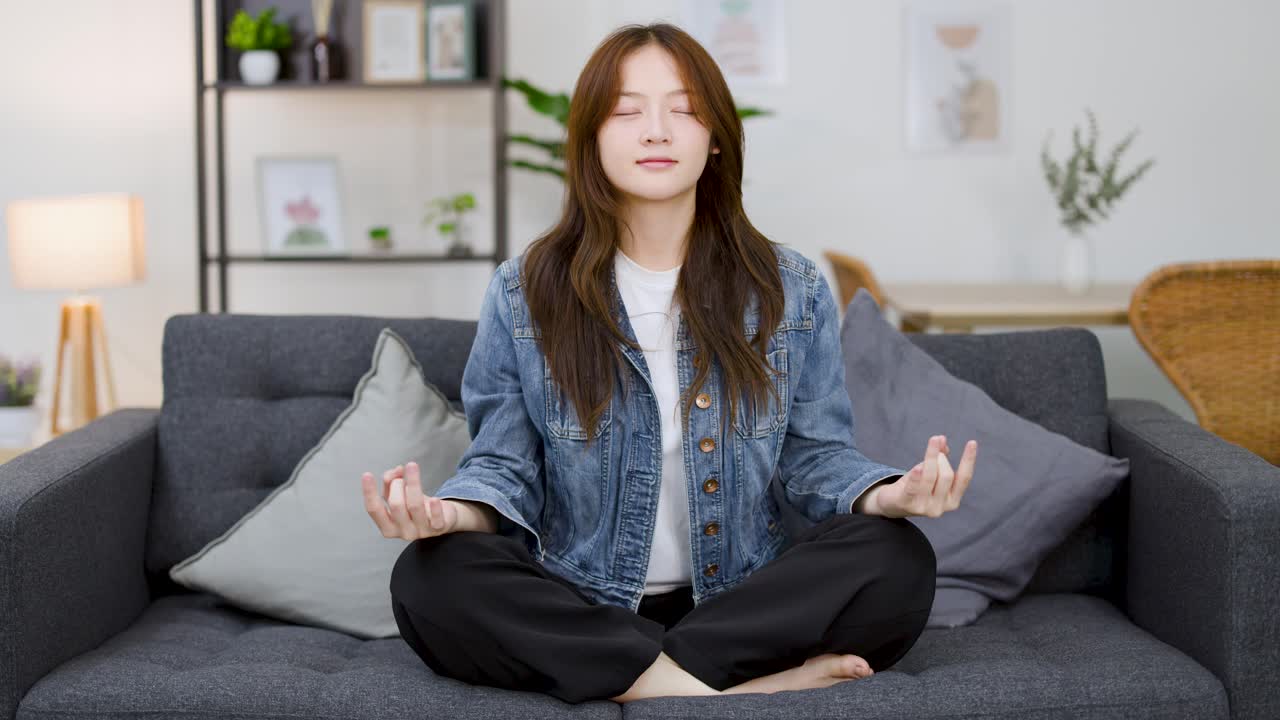 一位年輕的亞洲女性盤腿坐在沙發上，閉著眼睛練習冥想。