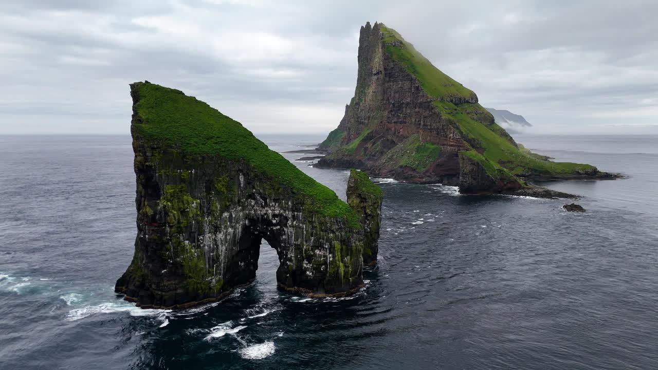 Cinematic aerial view of Drangarnir sea stacks rising dramatically from the Atlantic Ocean near Vágar, Faroe Islands, showcasing rugged cliffs, lush green slopes, and misty Nordic seascape