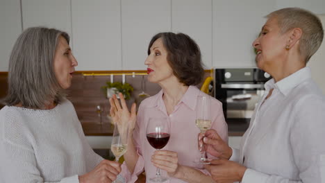 grupo de alegres amigas mayores hablando, riendo y bebiendo vino en la cocina 1
