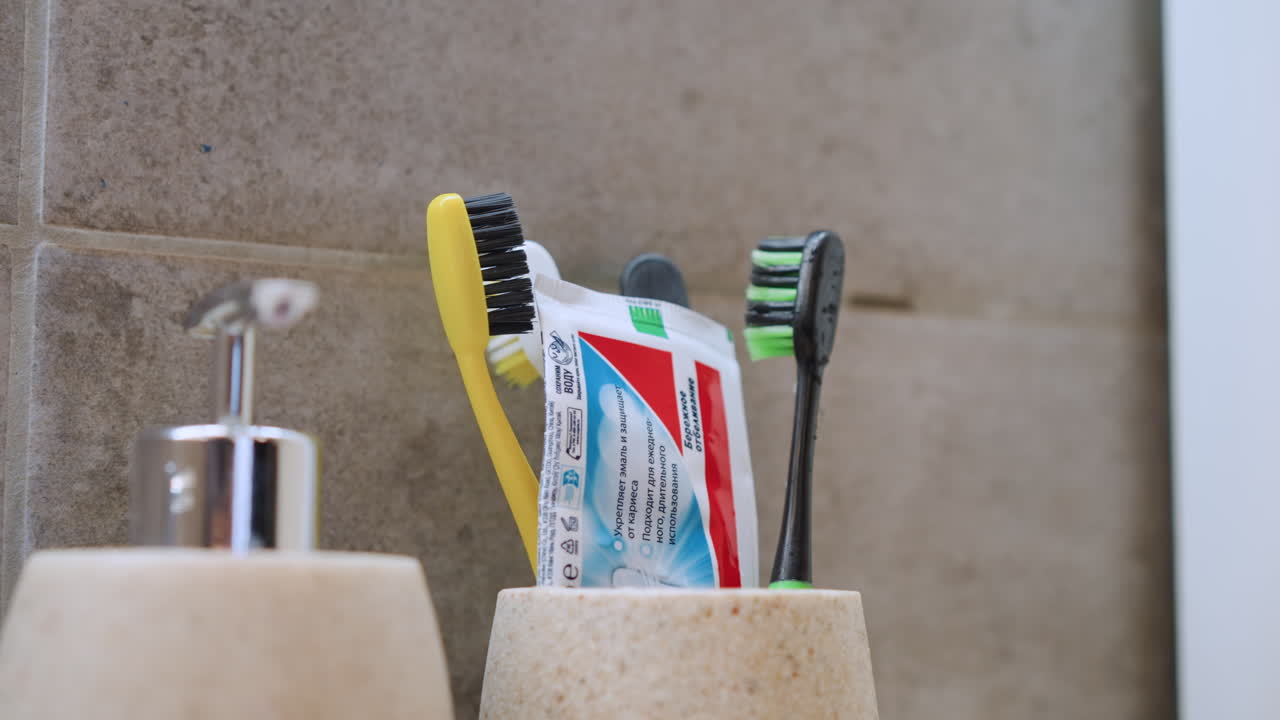 Toothbrushes and toothpaste in holder, closeup of two colorful brushes and squeezed paste tube beside soap dispenser on tiled bathroom wall, bright morning light, minimalist family routine, clean