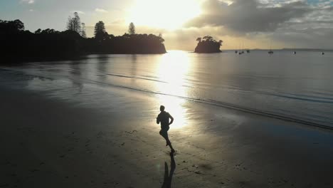 cinematic aerial tracking shot of silhouetted young man running on a beach in auckland, new zealand