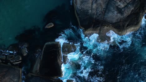 top down aerial view of waves crashing into boulders in the ocean