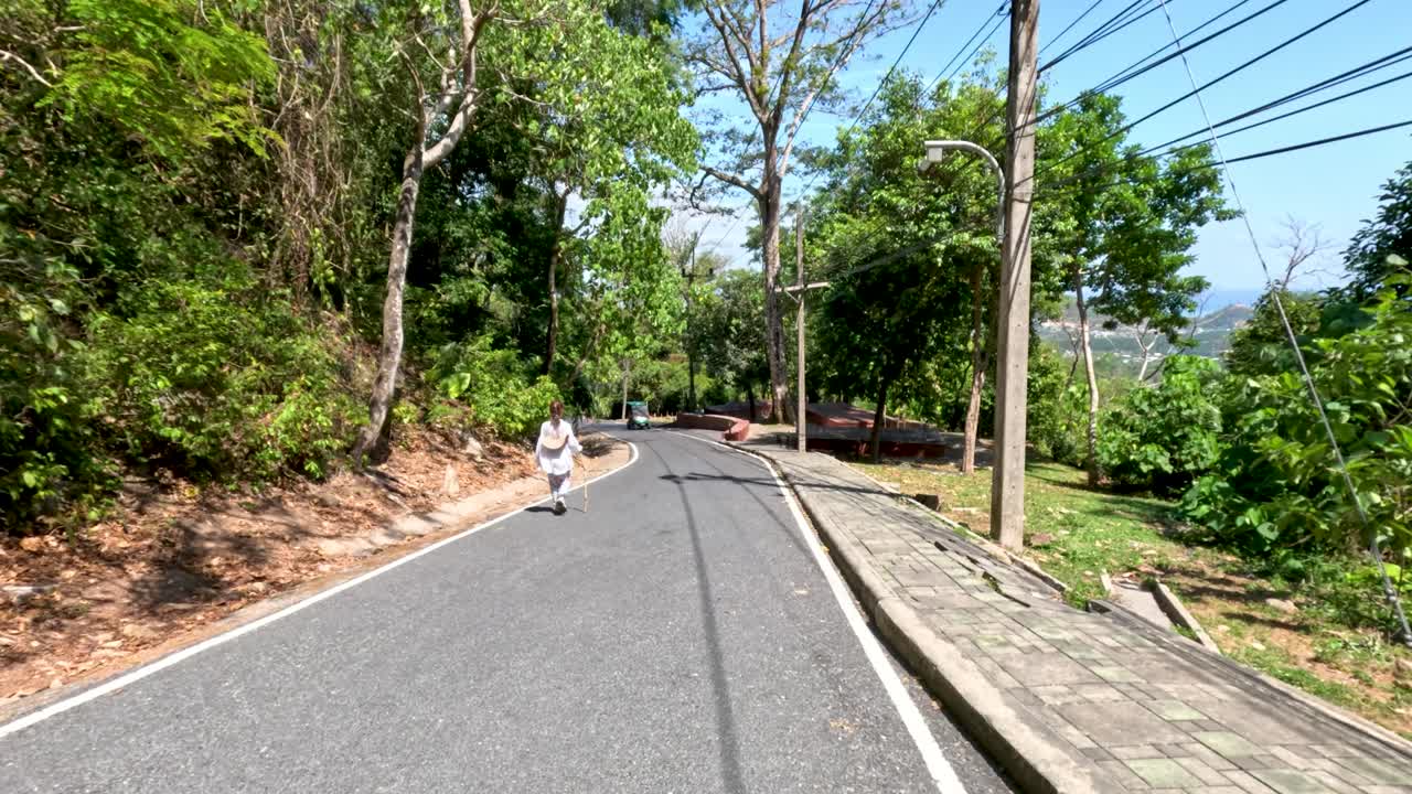 Individual jogs uphill on tree-lined road in bright daylight, camera slowly tracking forward
