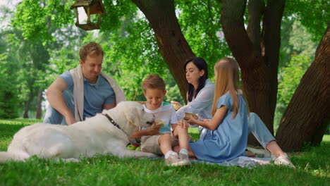 una familia feliz alimentando a un perro blanco en un picnic. la gente se divierte en la hierba verde.