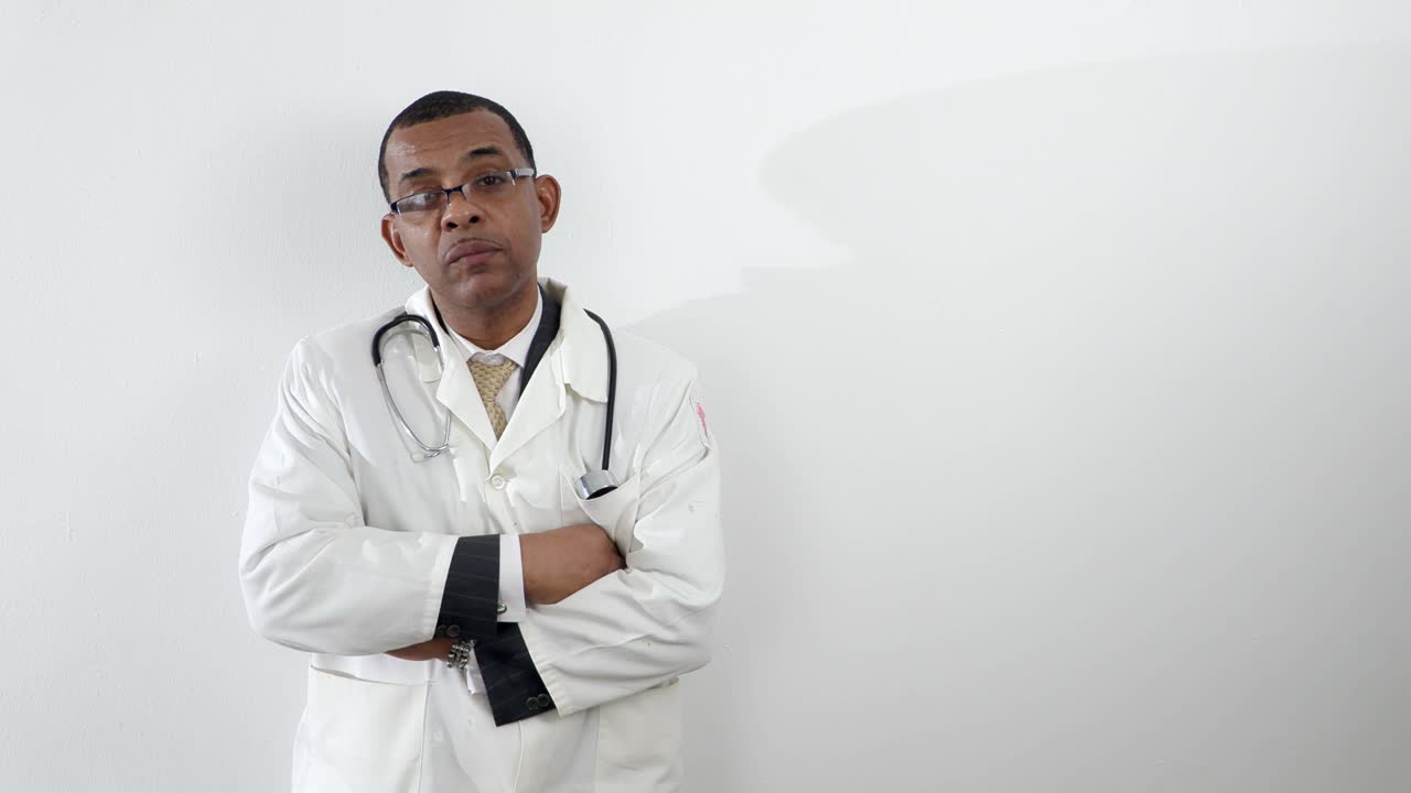 Jamaican African-American man doctor in white lab in glasses, coat turn his head to camera, serious tired look, against white wall background isolated mid shot