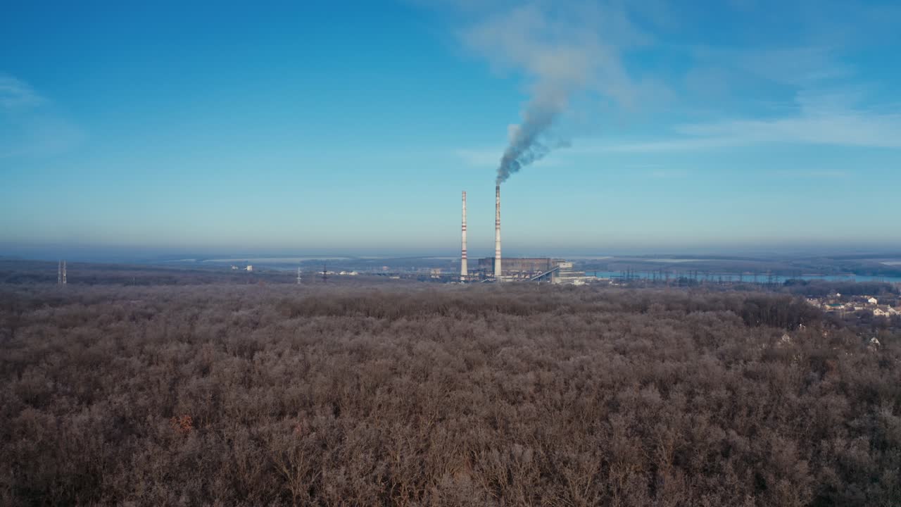 Flight over the forest to industrial factory. Top view of brown trees in winter. Manufacturing with two pipes among nature under blue sky. Aerial view.