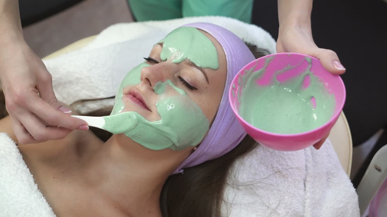 Facial mask on face. Therapist applying face mask to a beautiful young woman in spa using cosmetics brush