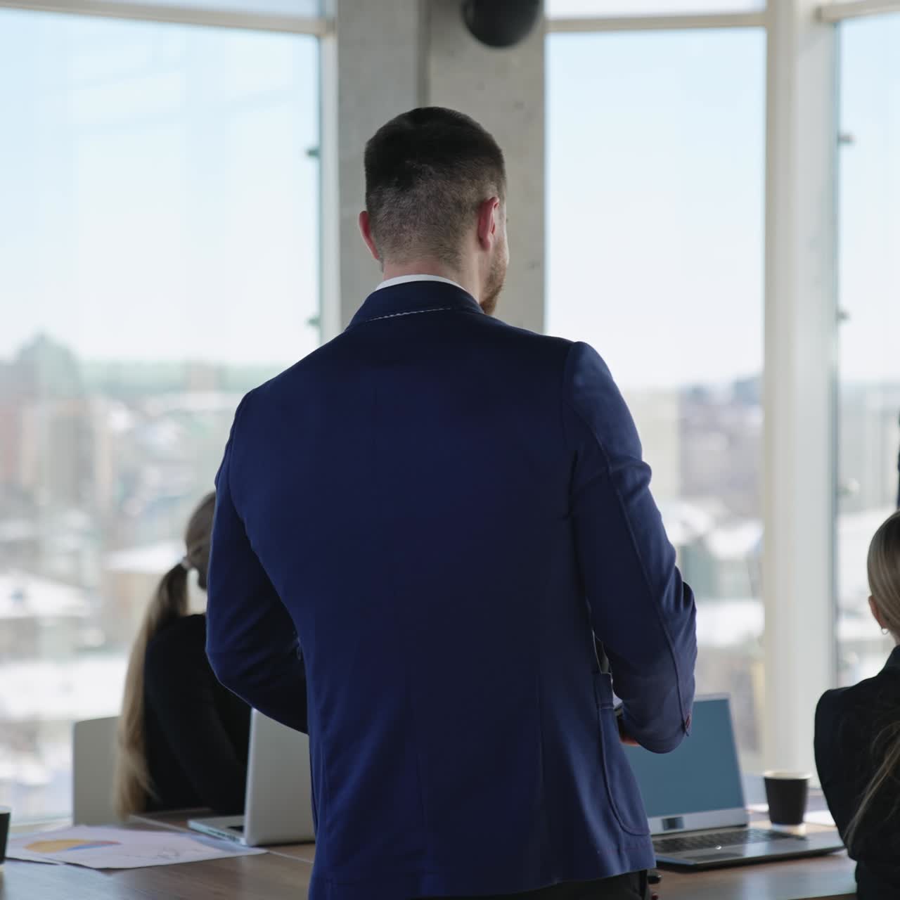 Handsome mature man in suit stands at foreground and then turns back and goes to the table. Team listening to their speaker at backdrop