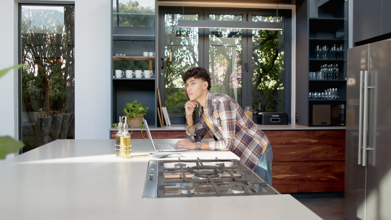Using laptop in modern kitchen, teenage boy leaning on counter, thinking