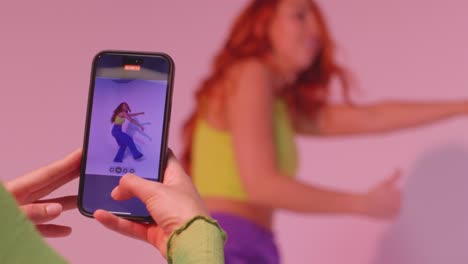 studio shot of woman taking photo of friend dancing on mobile phone against pink background 7