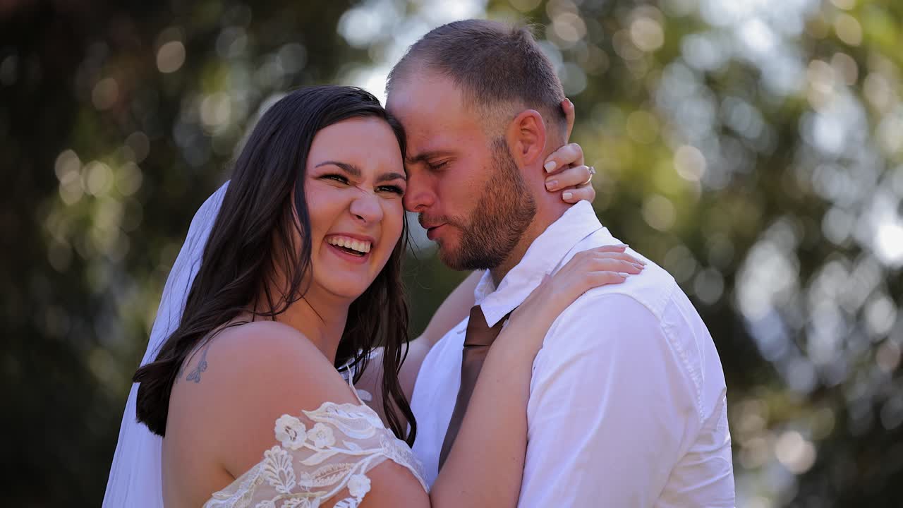 Noiva morena compartilha uma piada rápida e um sorriso com o marido em um jardim verde.