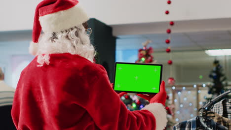 Christmas-ornate-shopping-mall-employee-impersonating-Santa-Claus-holding-mockup-tablet,-taking-a-break-from-job-to-watch-videos-on-chroma-key-screen-while-clients-walk-around,-close-up