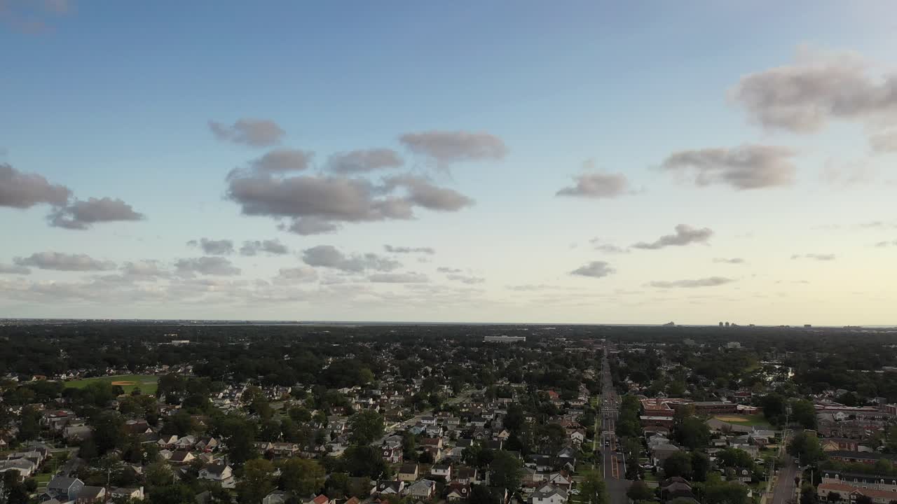 An aerial shot over a suburban, Long Island neighborhood. The drone is high enough to see the horizon. The camera slowly descend while tilting up. It is a sunny day with a few clouds in the blue sky