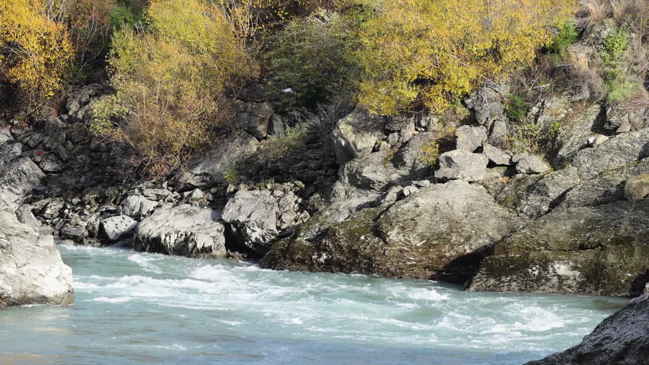 A fast-flowing river navigates rocky terrain in Queenstown, New Zealand, surrounded by autumn foliage under natural daylight