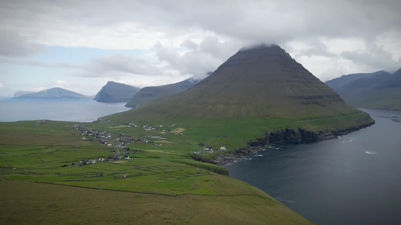 capturando una dramática vista aérea de viðareiði, las islas feroe, revelando un pueblo remoto anidado entre el mar y las altas montañas envueltas en nubes, una toma en órbita de avión no tripulado en cámara lenta
